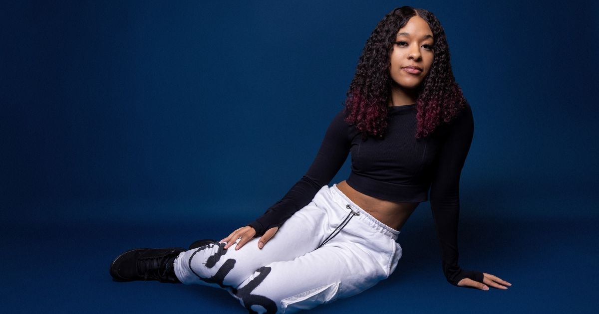 A Woman Poses In A Black Top And White Pants On A Blue Background For A Picture.