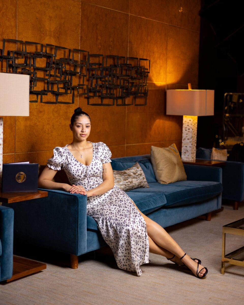 A Woman In A White Floral Dress Sits On A Blue Sofa In A Modern Lounge With Warm Lighting, Decorative Wall Art, And Red October Firm Lamps.