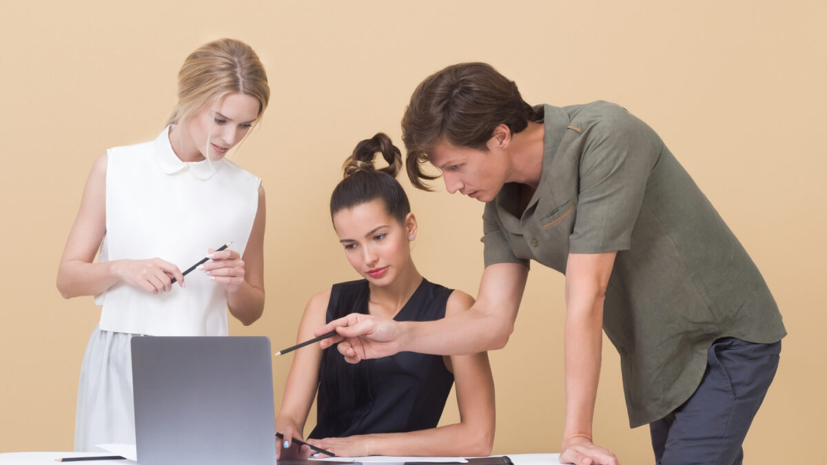 Three Business People Providing Feedback On Photos Displayed On A Laptop Against A Beige Background.