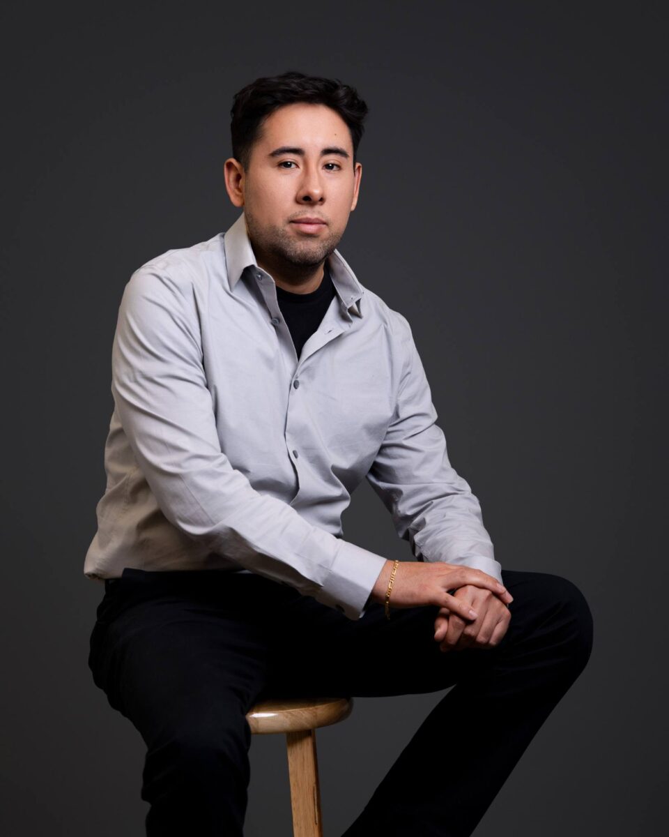 A Man In A Light Gray Button-Up Shirt And Black Pants Sits On A Wooden Stool Against A Plain Dark Background, Facing The Camera With Hands Resting On His Knee, Embodying The Understated Style Of The Red October Firm.