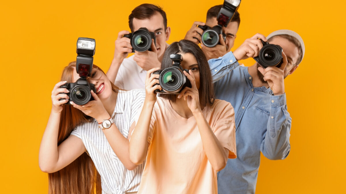 Individuals From Various Photography Groups Capturing Moments With Their Cameras Against A Vibrant Yellow Backdrop.