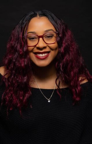 A Black Woman With Red Hair And Glasses Smiling.