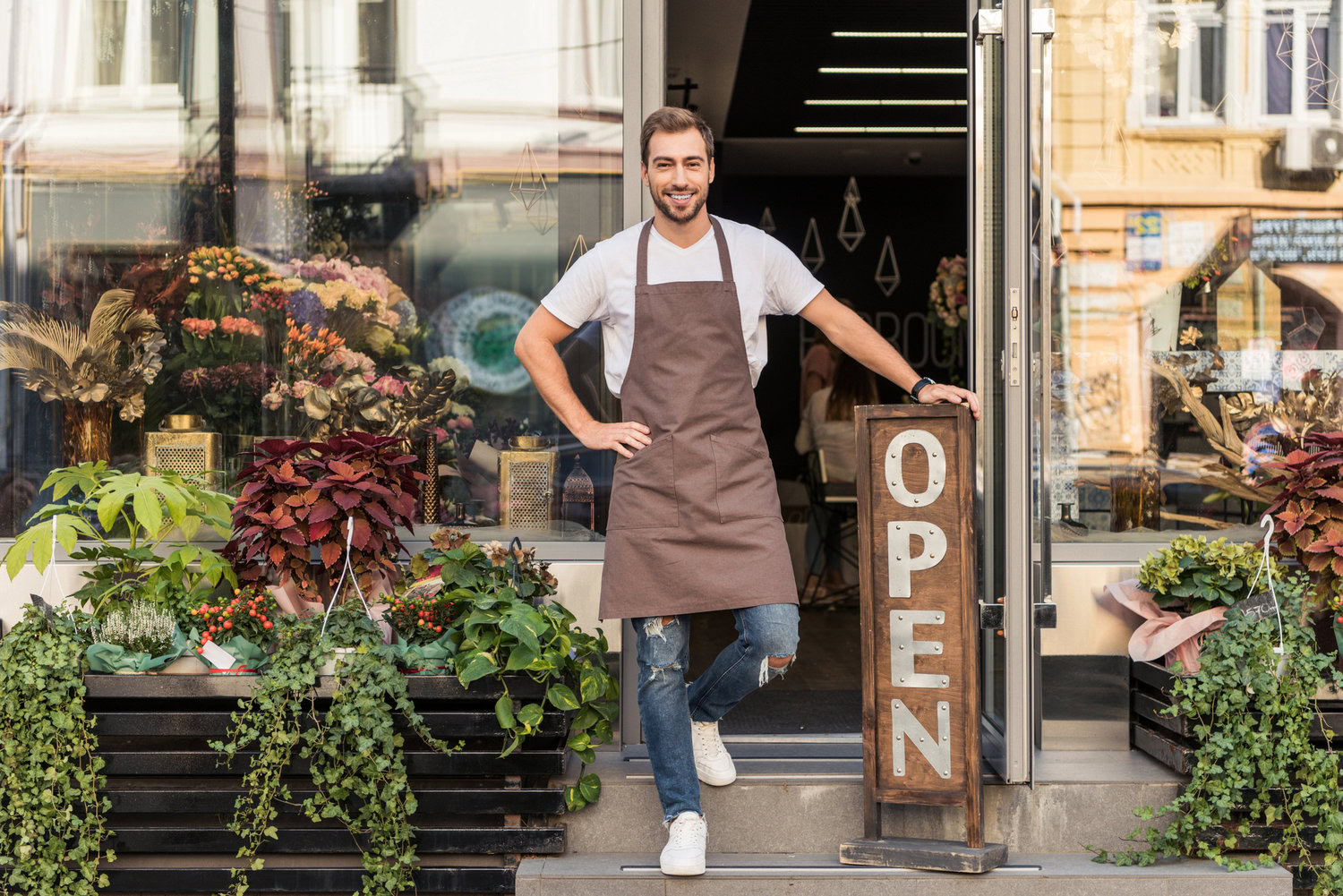 A Man Wearing An Apron Stands In The Doorway Of A Flower Shop With Plants And Flowers Around Him And A Sign That Reads &Quot;Open,&Quot; Offering His Services To All Who Enter.