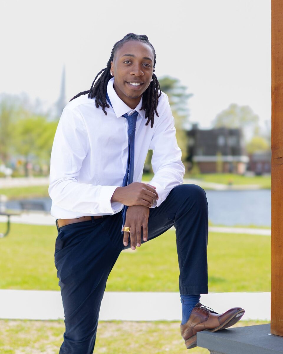 A Man In A White Shirt, Blue Tie, And Dark Pants Poses Outdoors With One Foot Raised On A Step, Smiling At The Camera—Capturing The Confident Spirit Of Red October Firm.