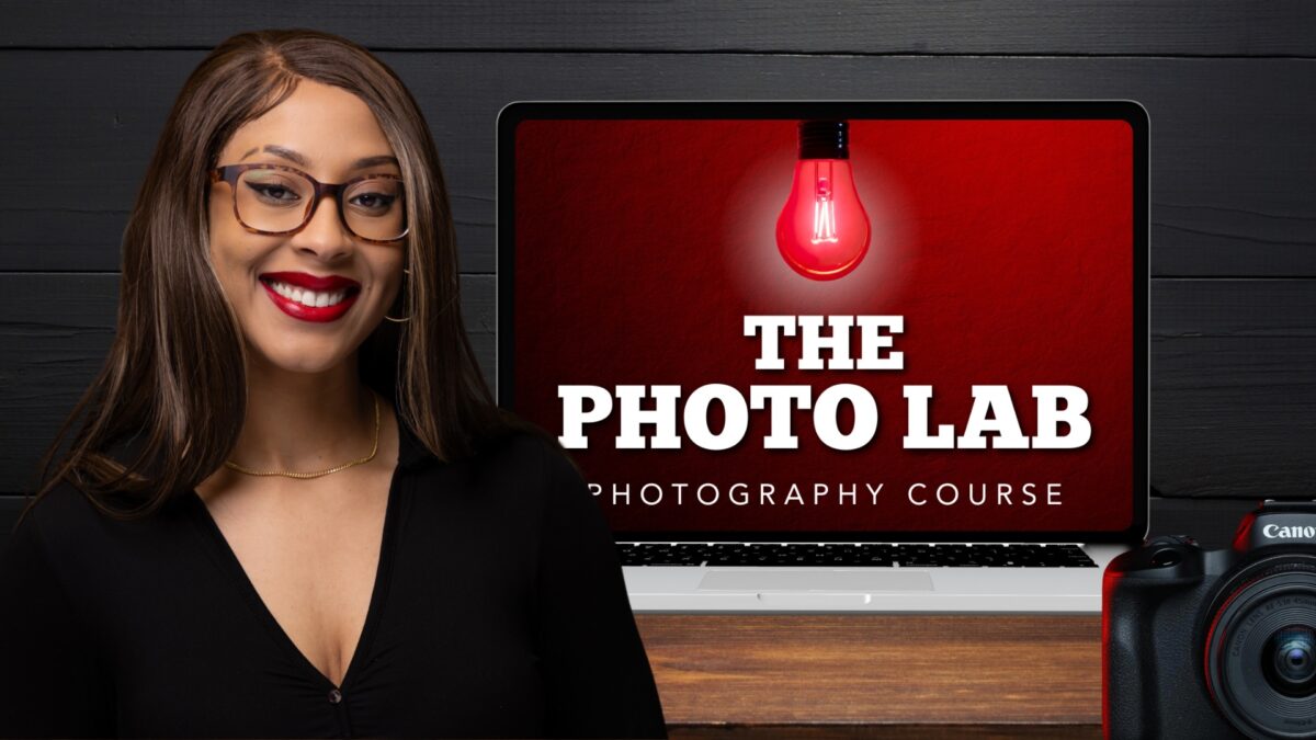 22 Ways To Market Your Photography Business For More Clients Smiling Woman With Glasses Facing The Camera, Standing In Front Of A Laptop Displaying &Quot;The Photo Lab Video Course&Quot; And A Red Light Bulb Image; Camera Partially Visible In The Foreground.