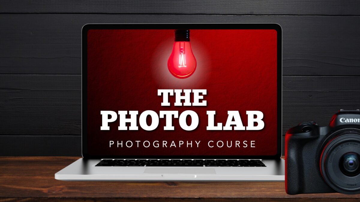 A Laptop Displaying &Quot;The Photo Lab Photography Course&Quot; And A Red Light Bulb On Its Screen. A Dslr Camera Is Positioned To The Right Of The Laptop.