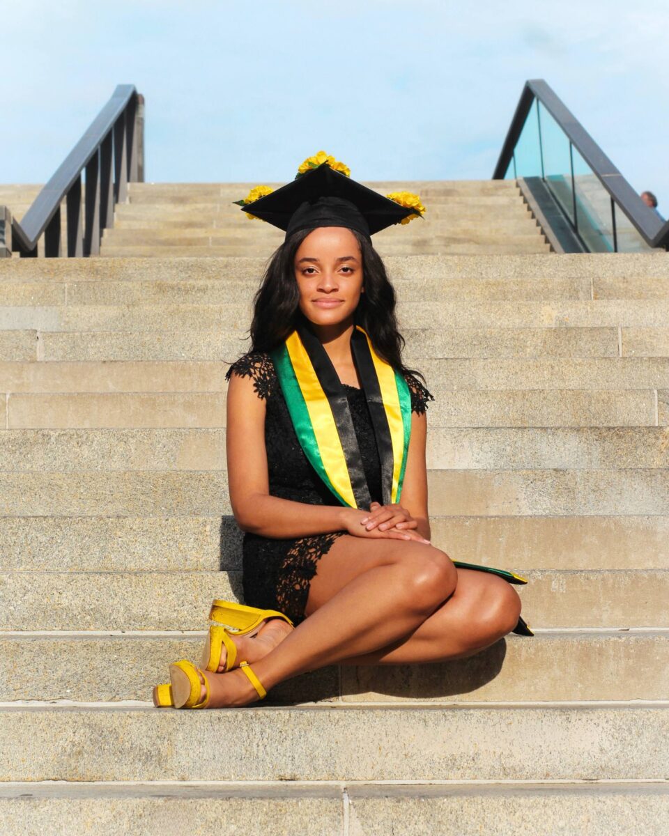 A Young Woman In A Black Dress And Graduation Cap, Representing Red October Firm, Sits On Outdoor Steps In A Green And Yellow Stole And Yellow Sandals, Looking At The Camera.