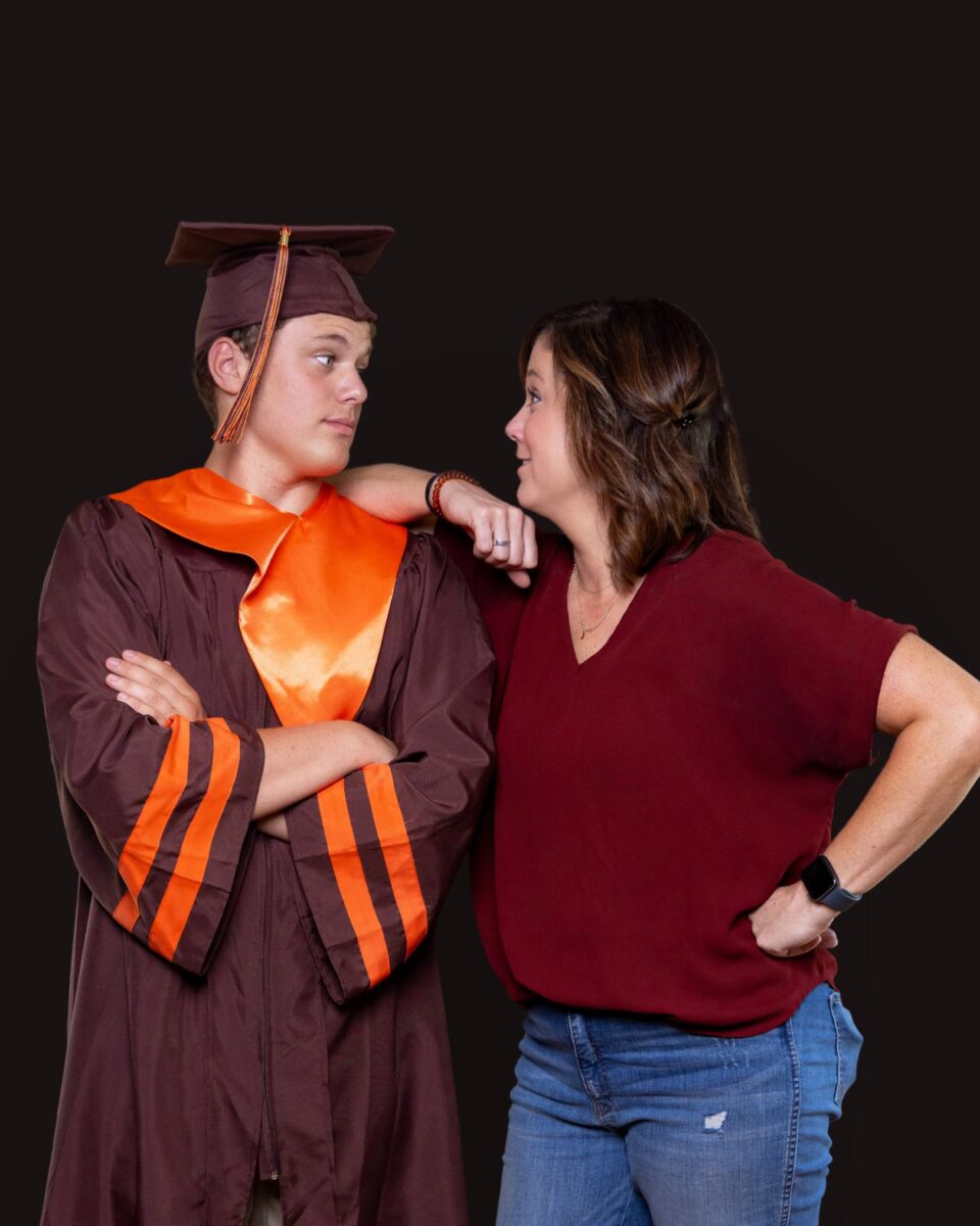 A Graduate In A Cap And Gown Stands With Arms Crossed While A Woman In A Casual Outfit, Embodying The Red October Firm Spirit, Leans On His Shoulder. Both Are Looking At Each Other Against A Plain Black Background.